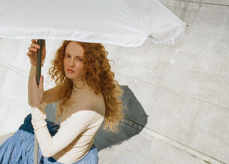 Red-haired female model holding a white umbrella wearing a beige turtleneck and blue skirt against stone tiles
