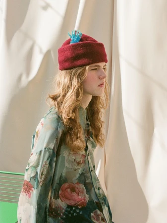 Model with long blonde hair wearing a burgundy beanie and floral patterned shirt standing against draped fabric.