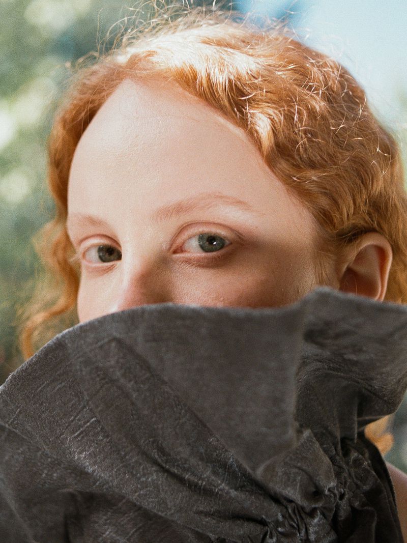 Close-up portrait of a woman with red hair holding up a dark textured collar to obscure the lower half of her face