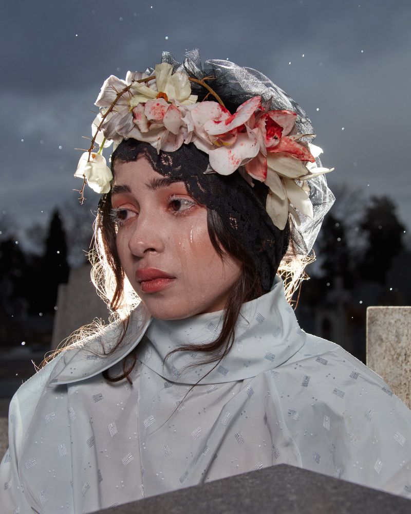 Young woman with floral headpiece and light blue coat standing in a cemetery with headstones visible on a cloudy day.