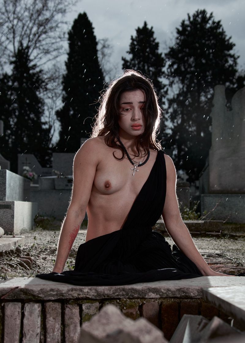 A topless young woman with necklace and black fabric stands in a graveyard with a tomb, gravestones and barren trees in the background.