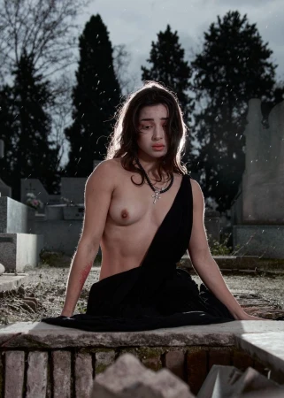 A topless young woman with necklace and black fabric stands in a graveyard with a tomb, gravestones and barren trees in the background.