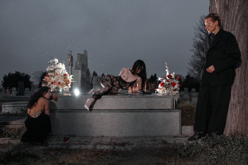 Three people in a cemetery at night: two women and a man, with tombstones, floral arrangements, and a somber, reflective atmosphere.