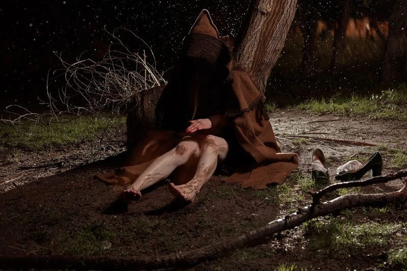 Person in brown hooded cloak sits by a tree in the dark night. Bare legs show caked mud; pair of high heels sit nearby on the grass.