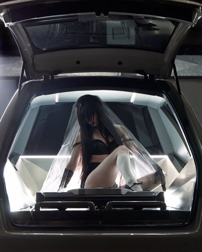 Woman with veil in the trunk of a black car posing in lingerie, illuminated by bright interior lighting, creating a striking contrast.