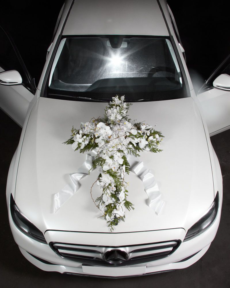 Front view of a white car decorated with a cross-shaped floral arrangement, featuring white flowers and ribbons against a black background.