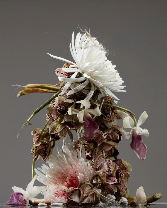 An intricate vertical sculpture composed of various orchids, white spider chrysanthemums, and stems against a grey backdrop.