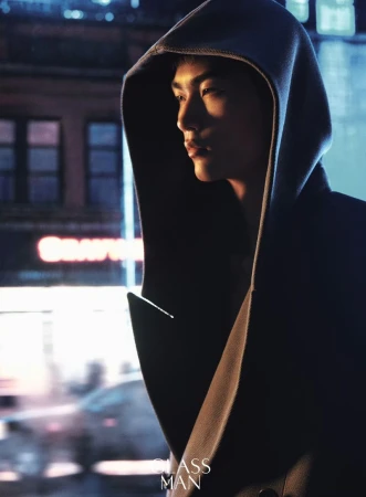 Man wearing a dark hooded coat standing outdoors at night with blurred urban lights in the background