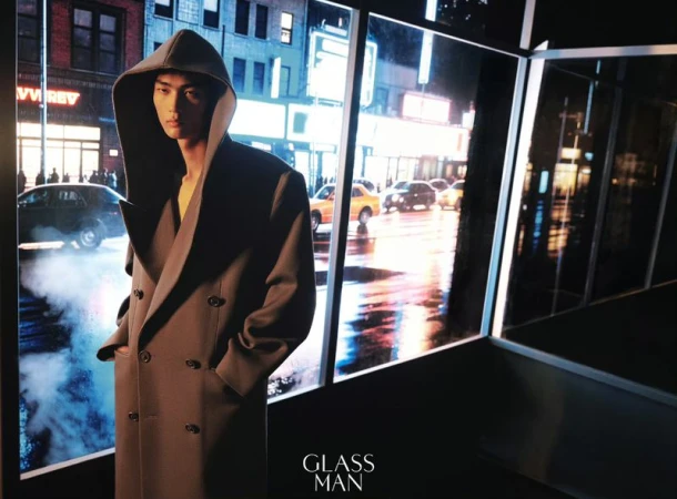 Model in a dark grey hooded coat standing indoors near large windows overlooking a wet city street at night