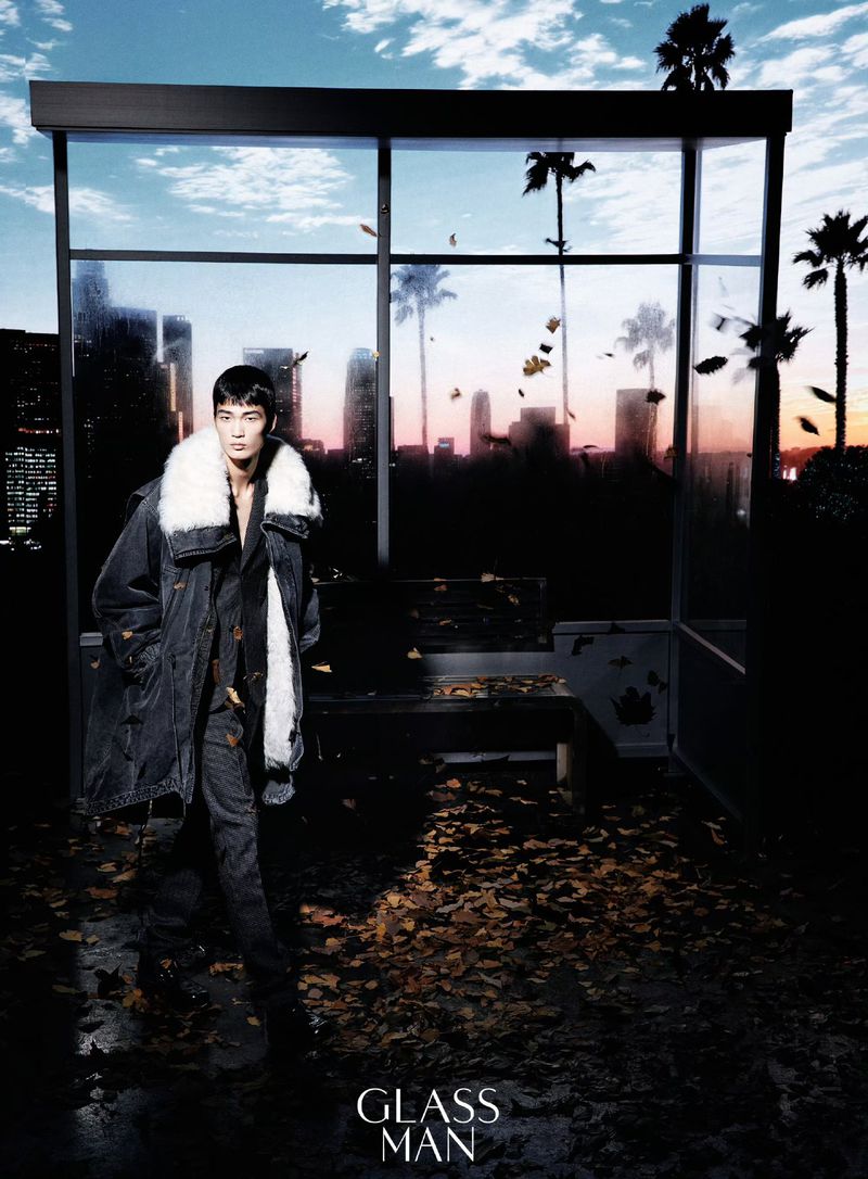Model wearing a dark coat with a white fur collar walking past a bus shelter with a sunset city skyline background
