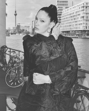 Monochrome portrait of Eartheater wearing a dark coat outdoors by a river with bicycles and cityscape