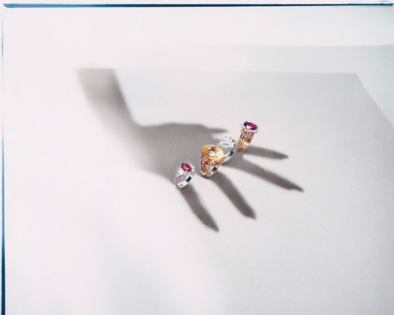 Four gemstone rings arranged in a row with a dramatic hand shadow cast across a pale neutral studio background.