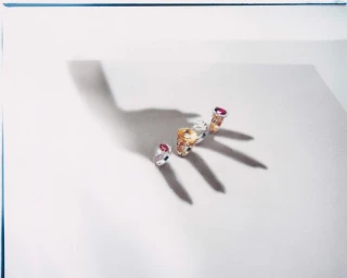 Four gemstone rings arranged in a row with a dramatic hand shadow cast across a pale neutral studio background.