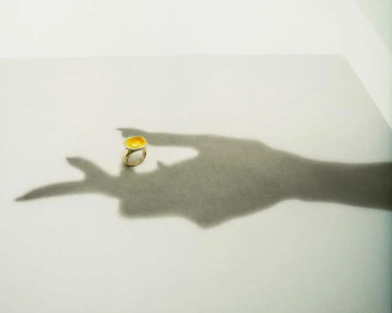 A gold ring with a yellow gemstone is placed on a white surface next to the dramatic shadow of a hand.