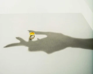 A gold ring with a yellow gemstone is placed on a white surface next to the dramatic shadow of a hand.