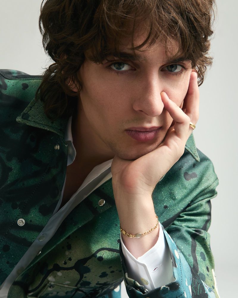 Close-up portrait of a man with wavy brown hair wearing a green printed jacket and gold bracelet, resting chin on hand against a white backdrop.