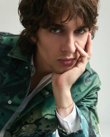Close-up portrait of a man with wavy brown hair wearing a green printed jacket and gold bracelet, resting chin on hand against a white backdrop.
