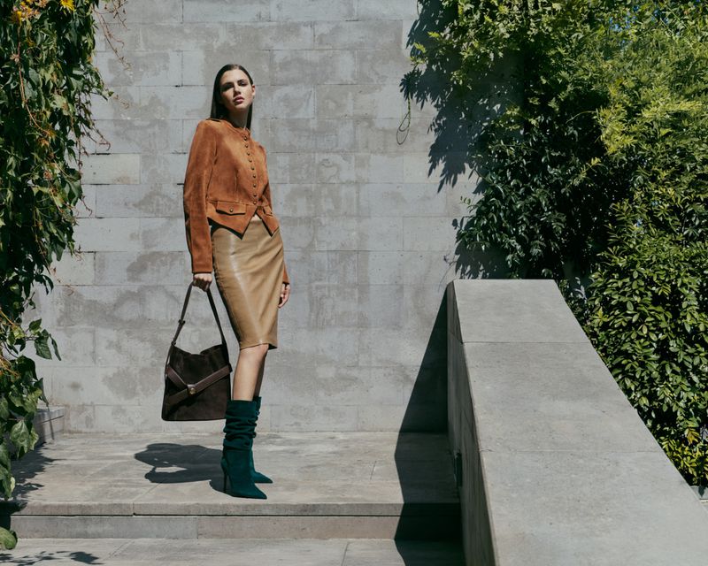 Model standing on stone steps wearing a brown jacket, tan skirt, and green boots while holding a dark shoulder bag.