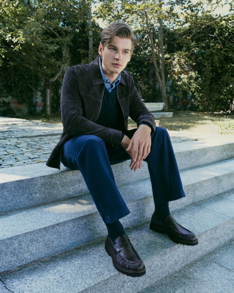 A man in a dark brown jacket and blue trousers seated on outdoor stone steps in an outdoor setting.