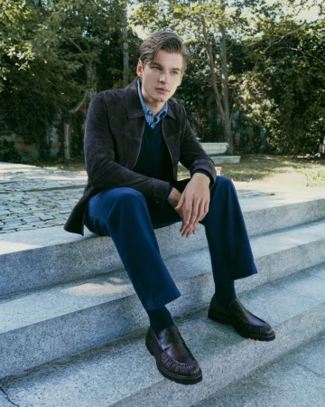 A man in a dark brown jacket and blue trousers seated on outdoor stone steps in an outdoor setting.