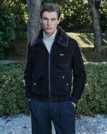 Male model wearing a dark brown suede bomber jacket with faux-fur collar and dark trousers outdoors in a garden setting.