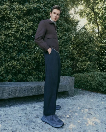 A male model wearing a dark brown jacket and trousers stands outdoors near a stone bench and lush green foliage.
