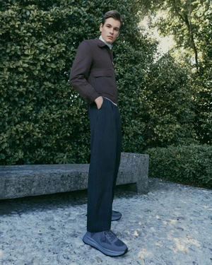 A male model wearing a dark brown jacket and trousers stands outdoors near a stone bench and lush green foliage.
