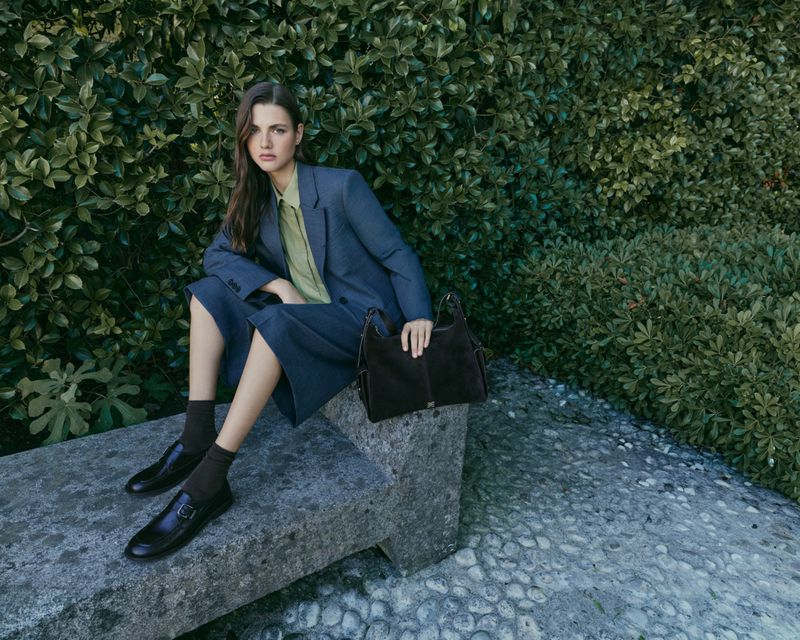 Woman in a grey suit jacket and culottes seated on a stone bench holding a dark brown handbag against a dense green foliage background