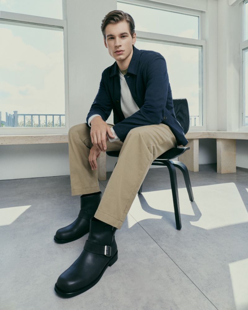 Male model seated on a black chair wearing a navy jacket, beige trousers, and black leather boots inside a bright room