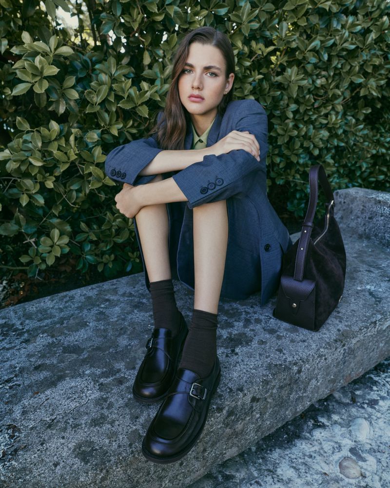 Model wearing blue suit jacket and dark loafers sitting on stone ledge in front of lush green foliage.
