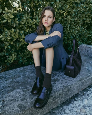 Model wearing blue suit jacket and dark loafers sitting on stone ledge in front of lush green foliage.