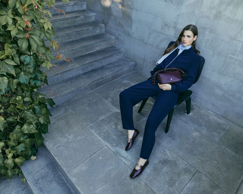 Woman in dark blue outfit and dark loafers seated on a chair outdoors near stone steps and climbing ivy.