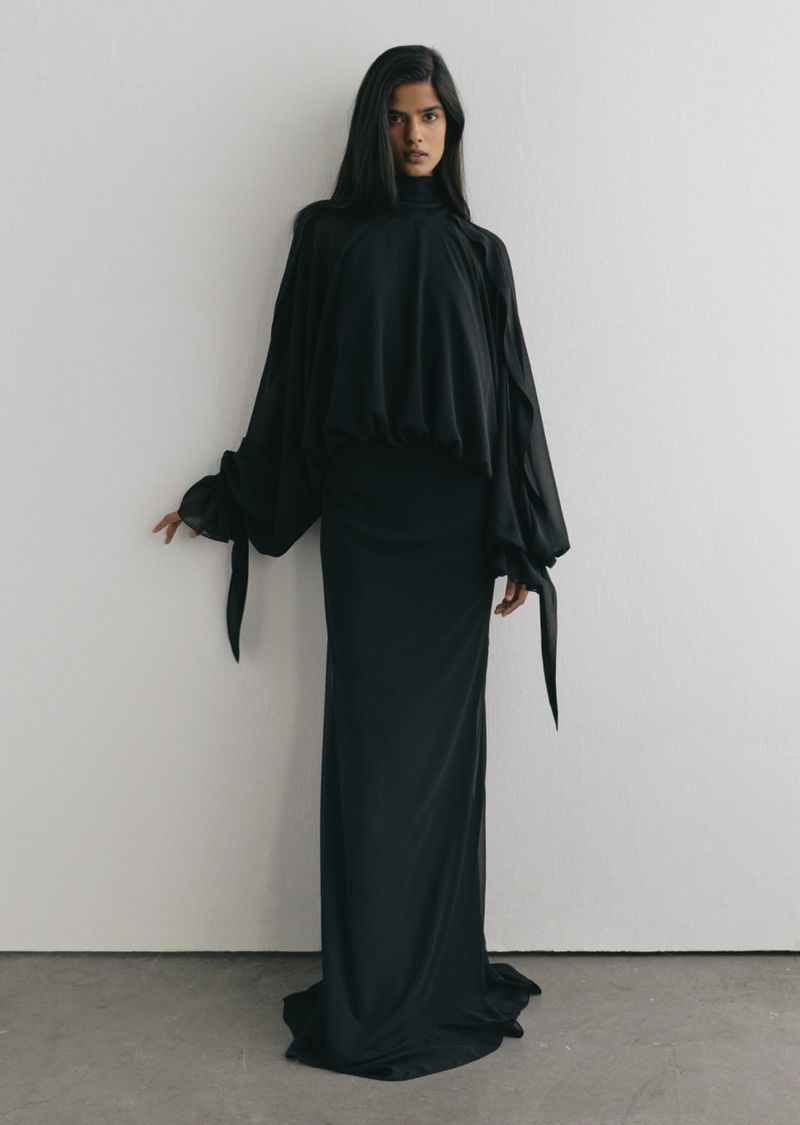 Model wearing a long black flowing dress with billowing sleeves against a neutral white background