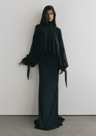 Model wearing a long black flowing dress with billowing sleeves against a neutral white background