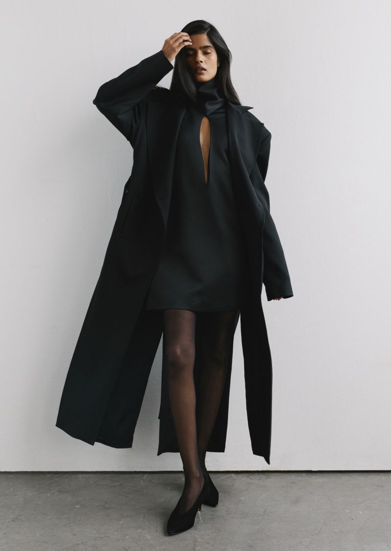 Model wearing a long black coat and dress standing against a plain light wall in a fashion lookbook image.