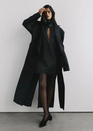 Model wearing a long black coat and dress standing against a plain light wall in a fashion lookbook image.
