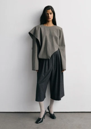 Model wearing a structured grey top and dark trousers against a plain white studio background.