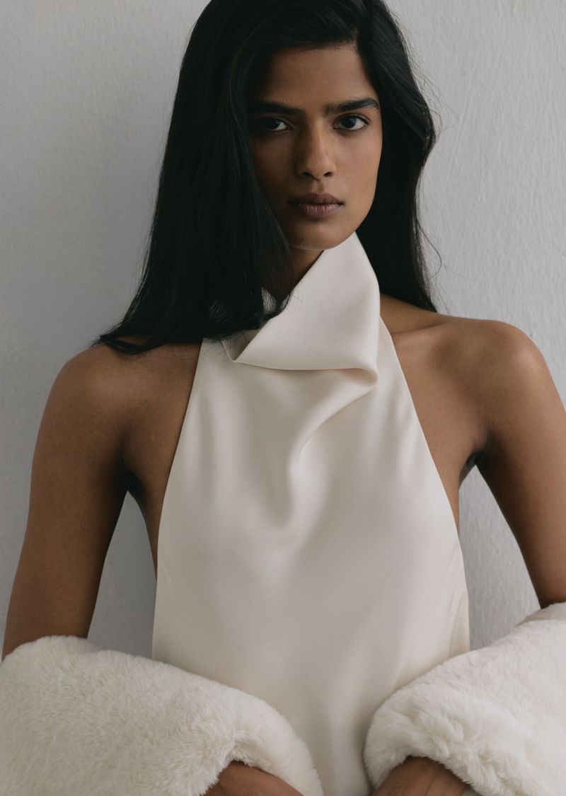 Close-up portrait of a woman wearing a cream halter top and white textured outerwear against a neutral grey wall.