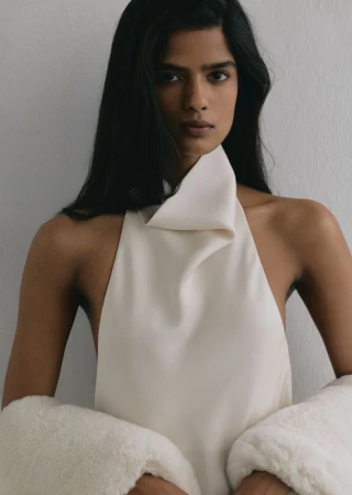 Close-up portrait of a woman wearing a cream halter top and white textured outerwear against a neutral grey wall.