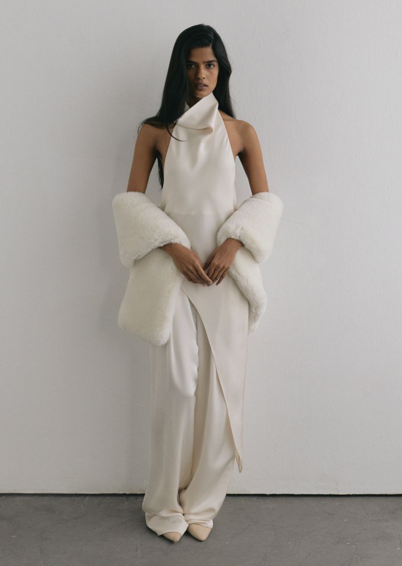 Woman wearing an ivory silk halterneck top and matching wide-leg trousers draped with a white faux fur wrap.
