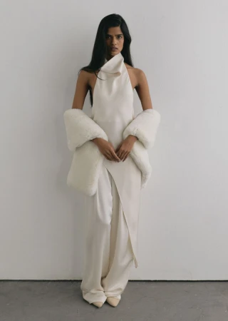 Woman wearing an ivory silk halterneck top and matching wide-leg trousers draped with a white faux fur wrap.