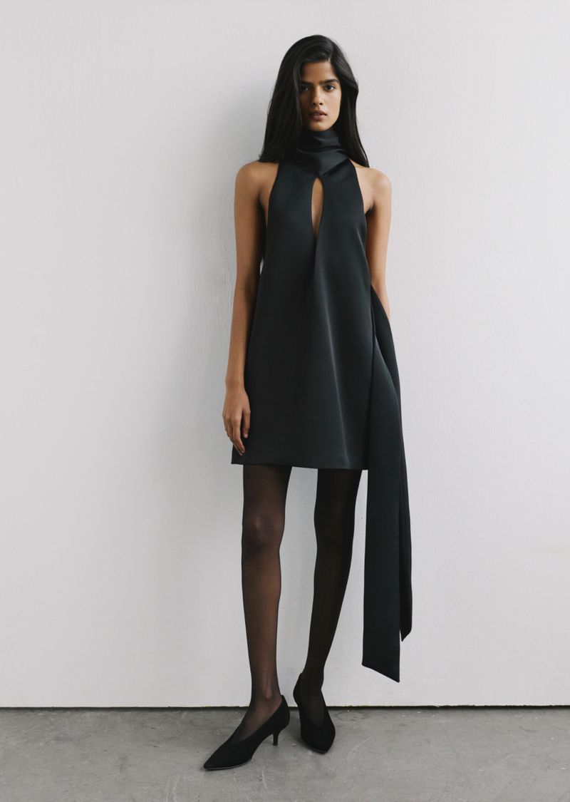 Model standing in a black halter mini dress with sheer black tights against a plain studio background.