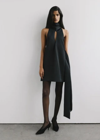 Model standing in a black halter mini dress with sheer black tights against a plain studio background.