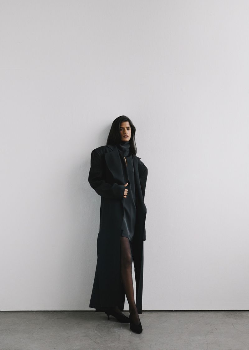 Model standing against a plain white wall wearing a long black coat and dark dress for MYBESFRIENDS FW26-27 collection.