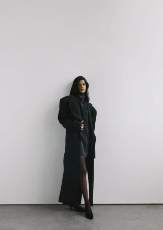 Model standing against a plain white wall wearing a long black coat and dark dress for MYBESFRIENDS FW26-27 collection.