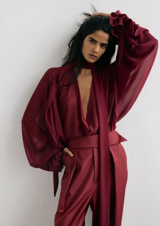 Model wearing a sheer burgundy blouse with billow sleeves and matching tailored trousers against a plain wall.