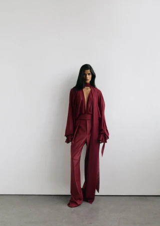 Model wearing a deep burgundy silk blouse and matching wide-leg trousers standing against a pale grey studio background.