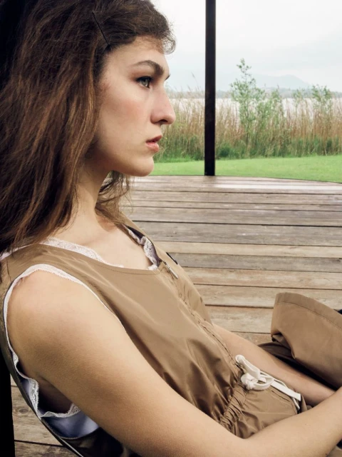 A young woman with long brown hair sits on a porch looking at a lake with grass and trees in the background. She wears a brown dress.