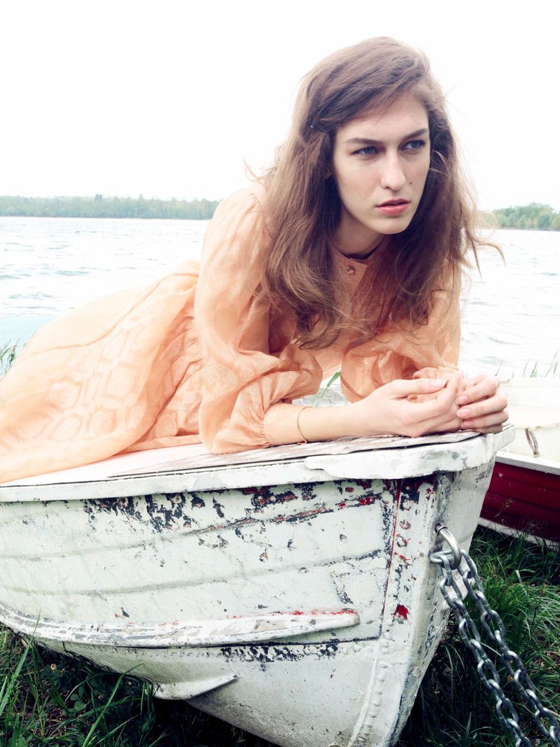 Woman in a peach dress resting on a white rowboat by a lake with trees under overcast sky. Fashion shot, outdoors, daytime.
