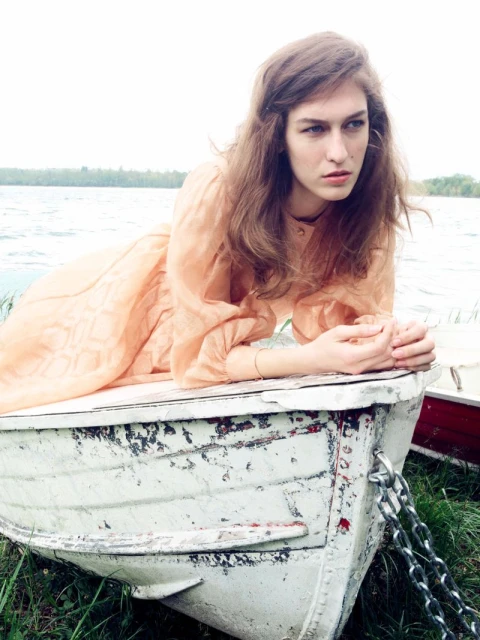 Woman in a peach dress resting on a white rowboat by a lake with trees under overcast sky. Fashion shot, outdoors, daytime.